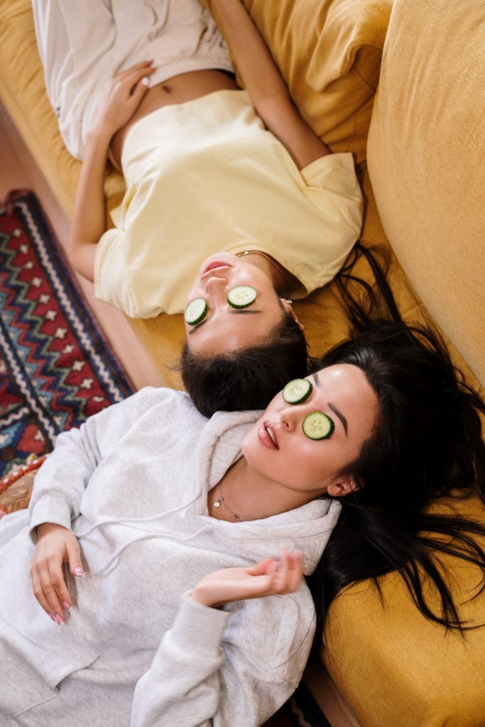 Two ladies lying against sofa having skincare routine with cucumber on eyes.